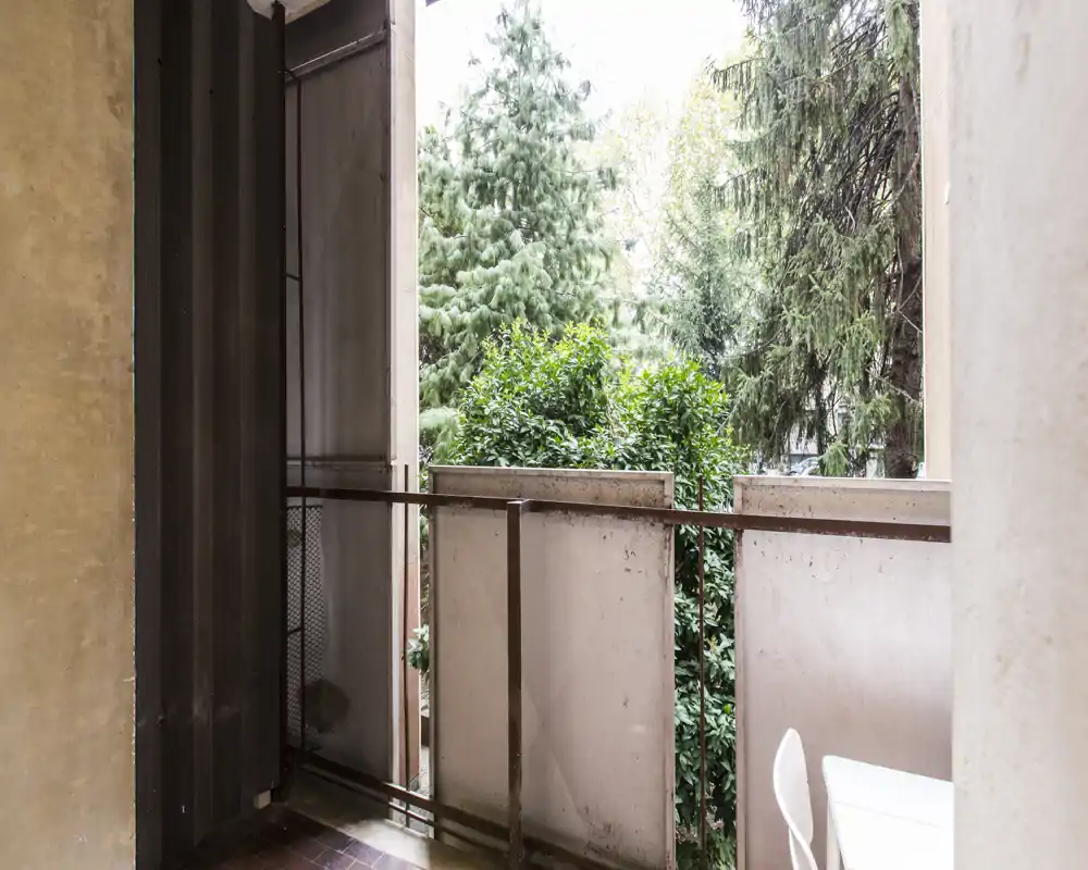 Piccolo balcone che guarda alberi maturi, con ringhiera in ferro semplice e seduta esterna parziale visibile; luce naturale luminosa e vegetazione gradevole.
