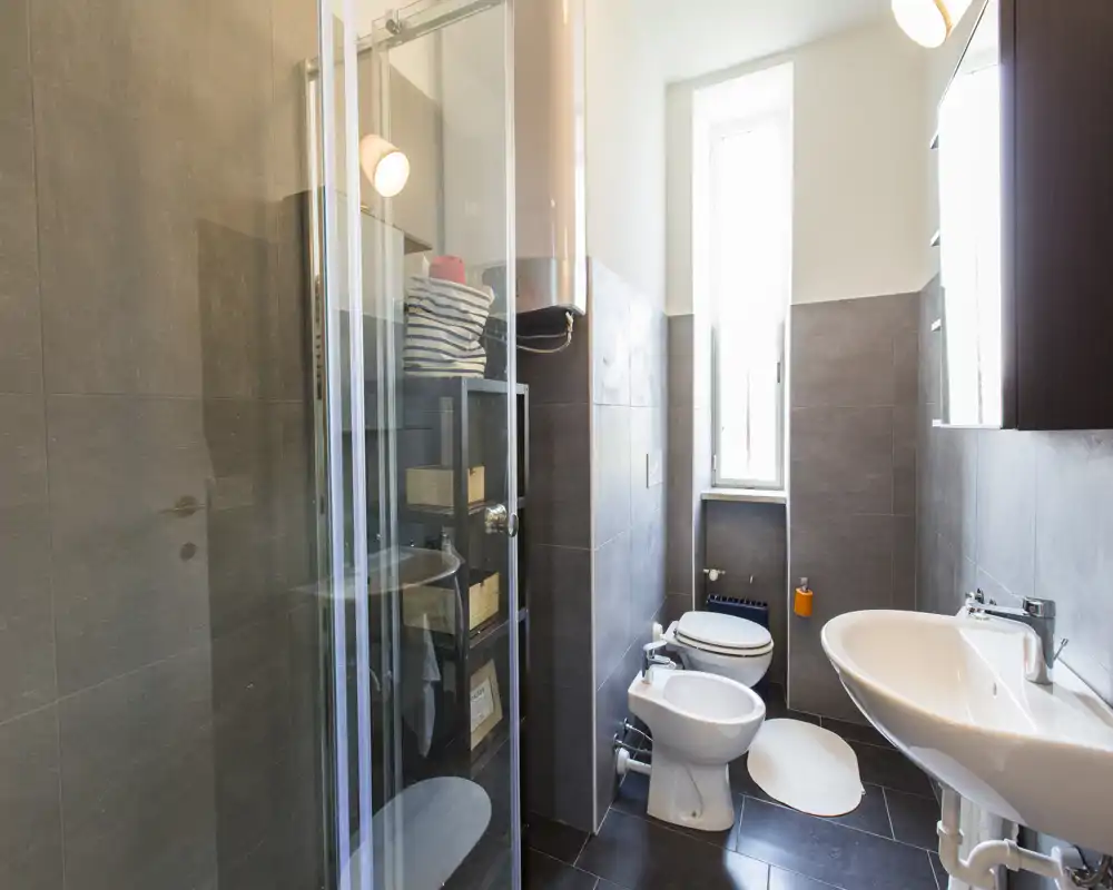 Bagno moderno e luminoso con doccia in vetro, bidet, WC e lavabo; piastrelle scure a contrasto con i sanitari bianchi e una finestra alta che garantisce luce naturale.
