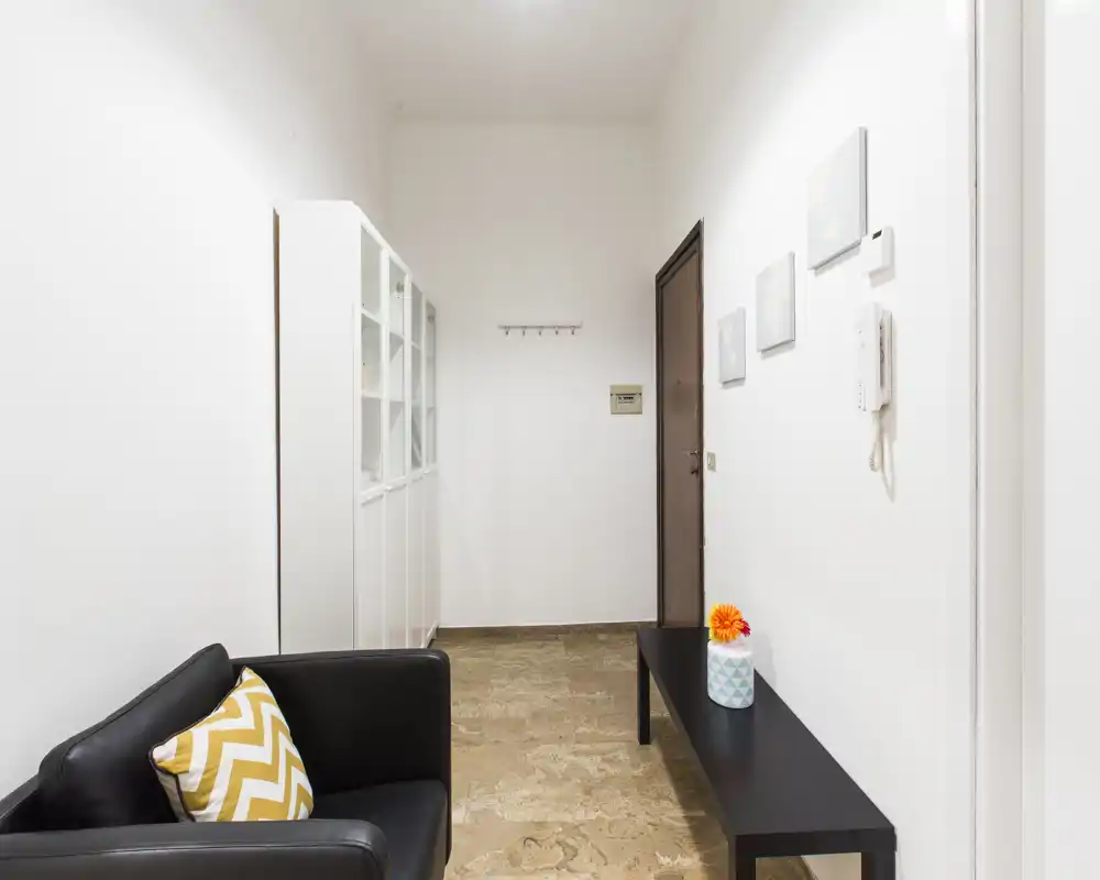 Ingresso luminoso e minimale con panca nera, appendiabiti e un piccolo angolo salotto — pulito, ben illuminato e accogliente per gli ospiti.