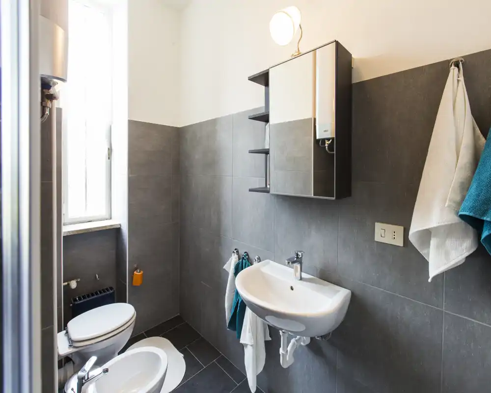 Bagno moderno e luminoso con piastrelle grigie, lavabo a parete, bidet e WC, pensile specchiato e ganci per gli asciugamani — pulito e funzionale.