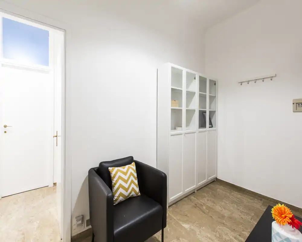 Piccola zona giorno con poltrona nera, cuscino a zigzag e armadietti bianchi; pareti neutre e buona luce naturale la rendono adatta a un angolo salotto o sala d'attesa.