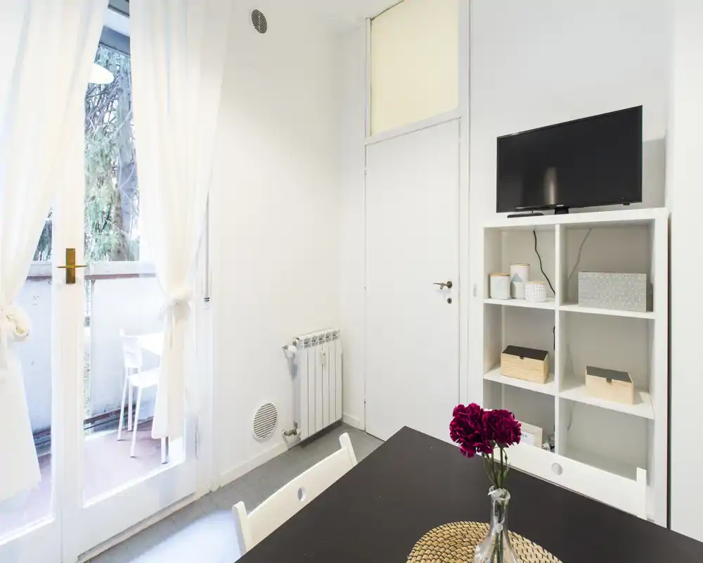 Angolo living/pranzo luminoso e ordinato con tavolo, sedie e scaffalatura; grandi porte a vetri si affacciano su un piccolo balcone e lasciano entrare luce naturale.