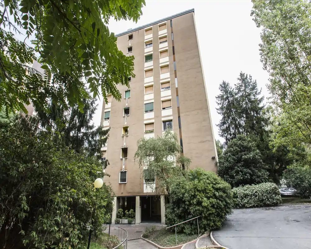 Vista esterna di un edificio residenziale con alberi circostanti e vialetto d'ingresso lastricato; si vedono la facciata, le finestre e il verde paesaggistico.