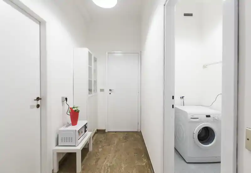 Bright, minimal entrance/hallway with white walls, a small bench holding a microwave and decorative plant, and a visible laundry room with a washing machine through an open door.