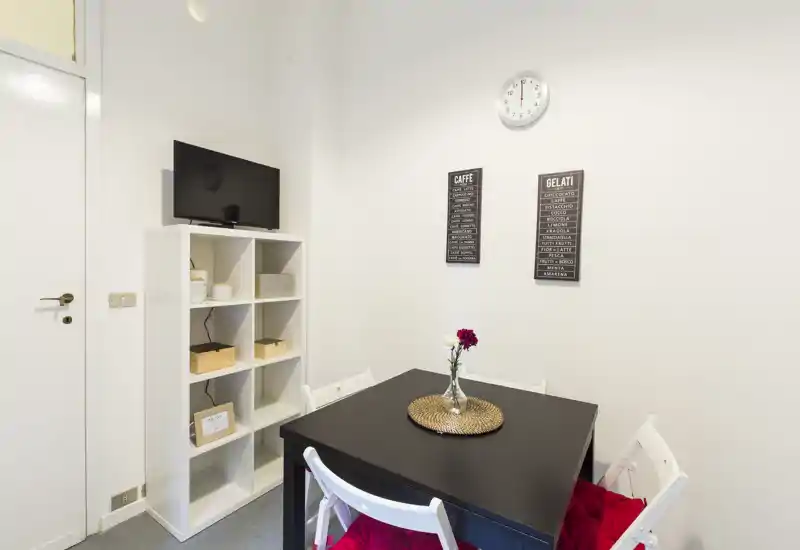 Compact dining corner with a black square table, four white folding chairs with red cushions, a decorative vase and shelving unit — bright, minimal and neatly styled for small-space dining.