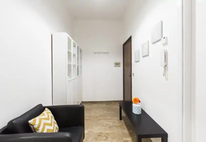 Bright, minimalist entry hallway with a black bench, coat hooks and a small seating area — clean, well lit and welcoming for guests.