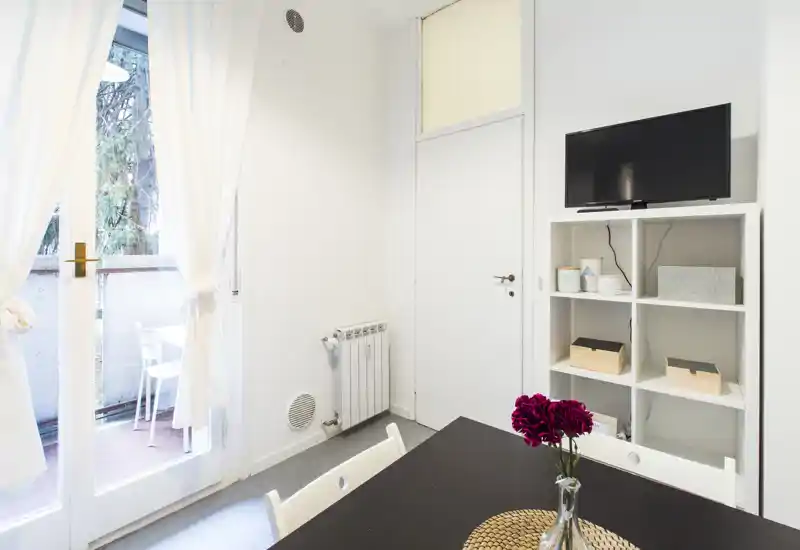 A bright, tidy living/dining corner with a table, chairs and shelving; large glass doors open to a small balcony letting in natural light.