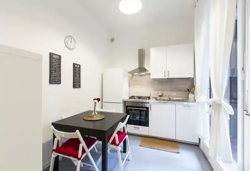 Compact modern kitchen with white cabinets, gas stove, oven and a small dining table by a bright window — neatly staged with cushions and a vase for a welcoming feel.