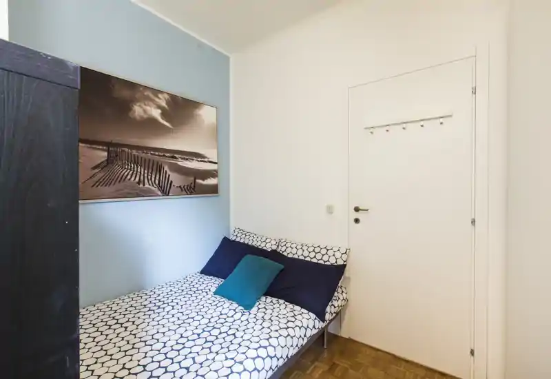 Compact, tidy bedroom with patterned bedding, two dark pillows and a teal cushion. Soft daylight and coastal wall art create a calm, welcoming atmosphere.