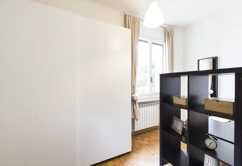 Bright, minimal bedroom corner with a large white wardrobe, parquet floor and an open shelving divider; natural light comes through the window creating a fresh, functional atmosphere.
