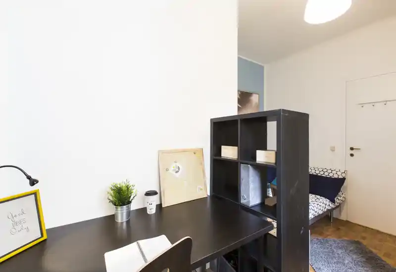Compact bedroom corner with a desk and partial single bed. The workspace features a dark desk, shelving divider, small plant and soft patterned bedding visible at the side.