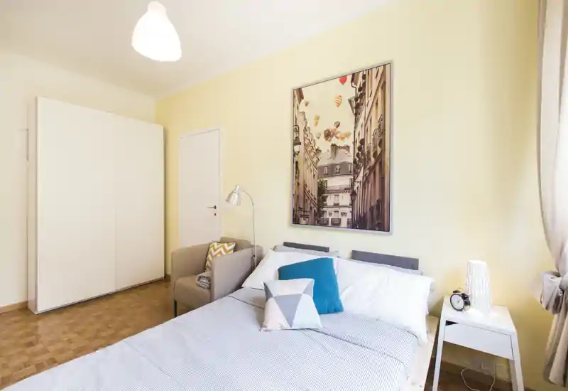 Bright, well-staged bedroom with a double bed, decorative pillows, bedside table and a comfortable armchair. Large wardrobe and framed artwork give a cozy, modern feel — ideal for rental photos.
