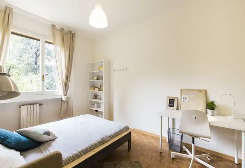 Bright, simply styled bedroom with a single bed, bedside cushions, shelving and a study desk by the window letting in abundant natural light.