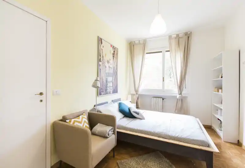 Bright, well-styled bedroom with a comfortable double bed, soft textiles and a cozy armchair by the door. Large window provides abundant natural light and neutral decor creates a calm atmosphere ideal for guests.