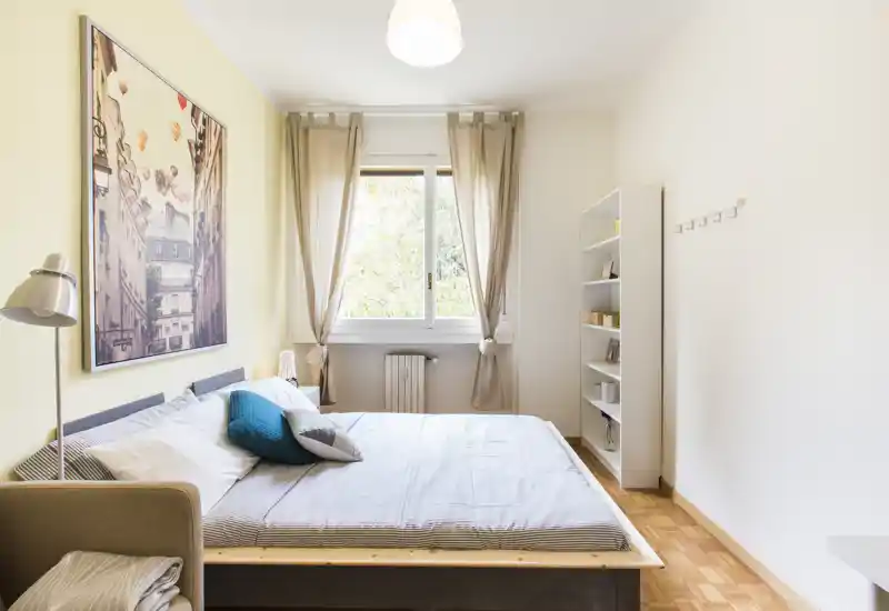 Bright, neatly styled bedroom with a large double bed, decorative cushions, a framed artwork and sheer curtains letting in natural light.