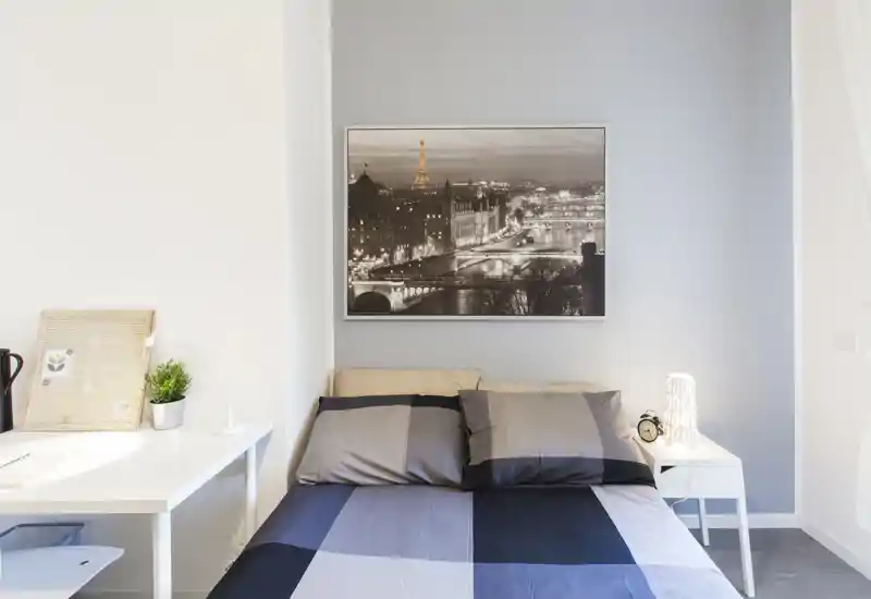 Bright, modern bedroom with a neatly made double bed, bedside table with lamp and an elegant cityscape print above the headboard. Clean lines, neutral palette and natural light create a calm, welcoming space ideal for a restful stay.