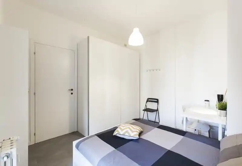 Bright, minimalist bedroom with a double bed dressed in a geometric duvet, large white wardrobe and a compact desk — ideal for a tidy short-stay rental.
