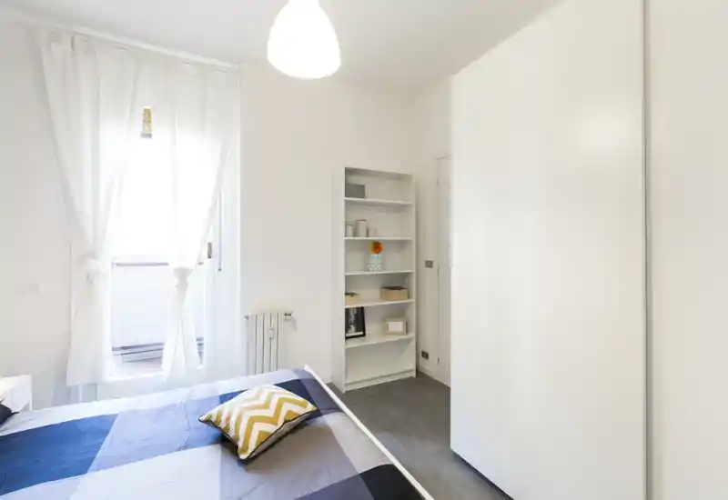 Bright, minimal bedroom with a neatly made bed, decorative cushion and a built-in wardrobe; abundant natural light from the window makes the space feel airy.