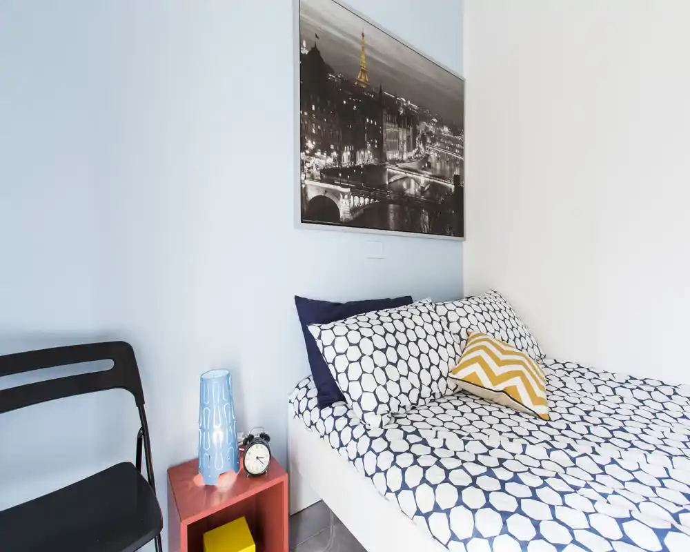 Angolo camera compatto e arredato con letto matrimoniale dal motivo geometrico, cuscini decorativi e una stampa della città sopra; moderno, luminoso e curato.