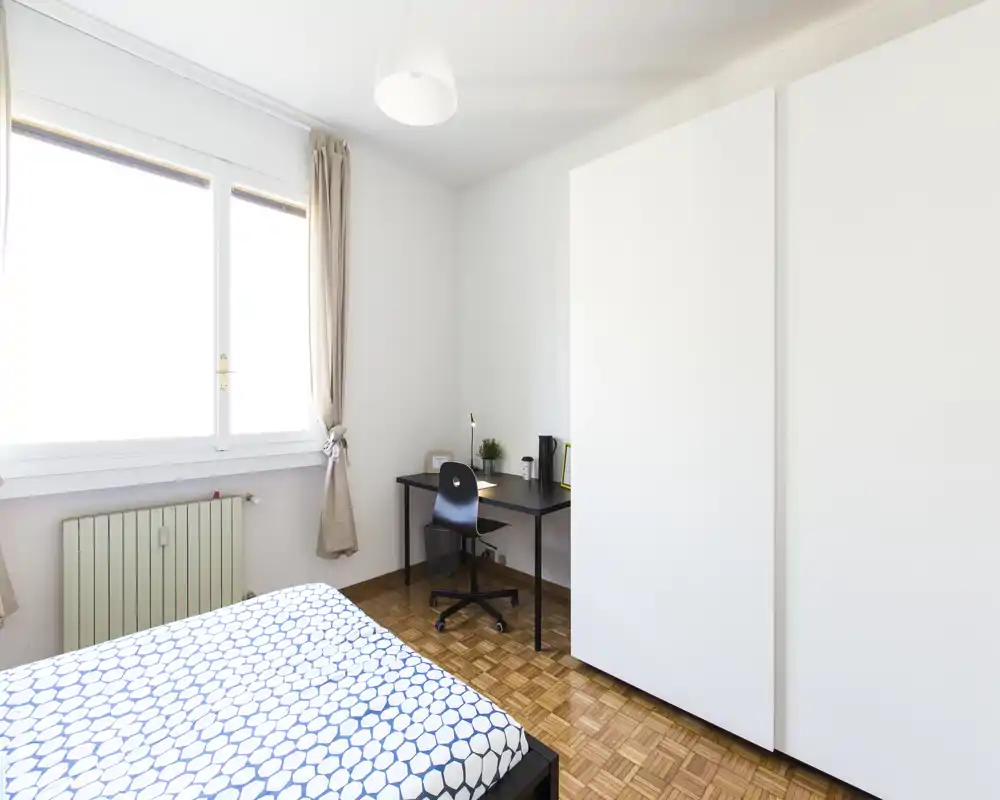Camera da letto luminosa e minimale con letto matrimoniale a fantasia, ampia finestra, scrivania compatta e armadio — ideale per un affitto in città ordinato.