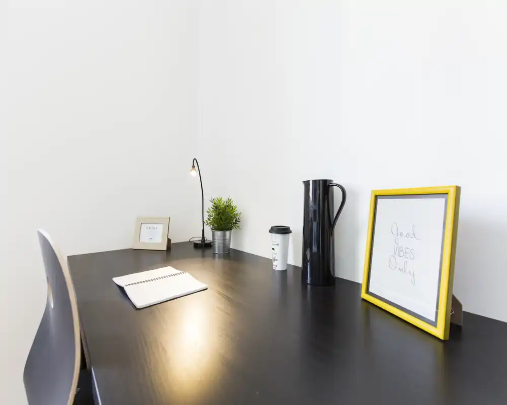 Angolo di lavoro luminoso e minimal con scrivania in legno scuro, piccola lampada, pianta e cornice: spazio ordinato ideale per concentrarsi.