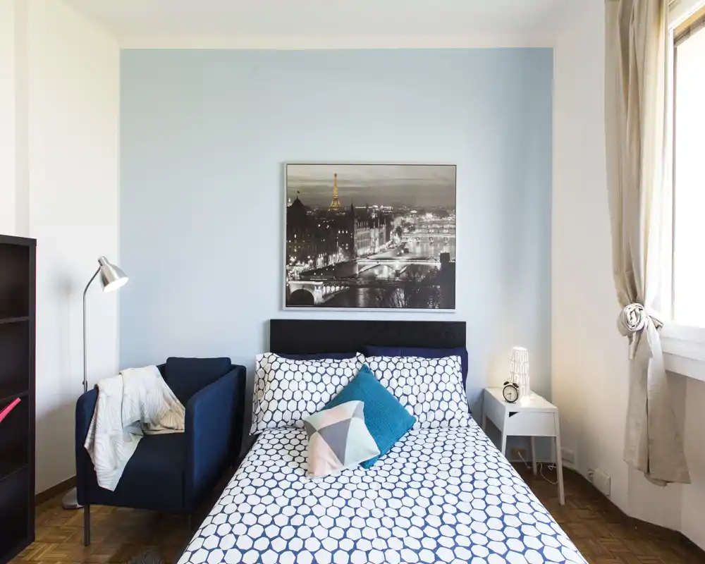 Camera da letto luminosa e ben arredata con letto matrimoniale con biancheria a motif, cuscini decorativi, comodino e una poltrona; ampia finestra che illumina l'ambiente.