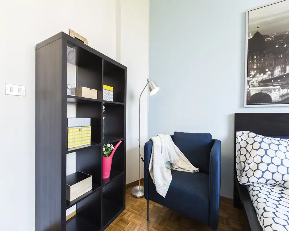 Angolo di camera moderno e luminoso con una libreria, una poltrona comoda con plaid e il letto parzialmente visibile con cuscini a motivo — ambiente ordinato e ben illuminato.