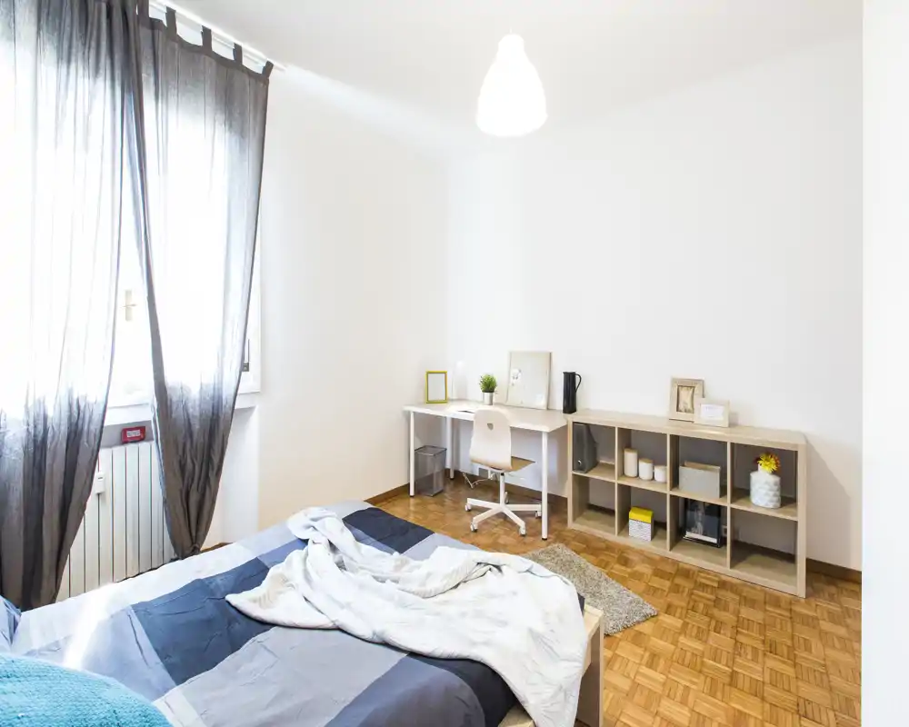 Bright, minimal bedroom with a double bed in the foreground, desk and shelving along the wall, and soft natural light from a large window.