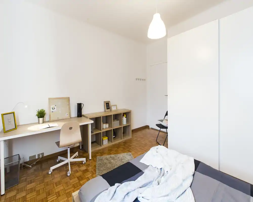 Bright, minimal bedroom with a neatly made bed in the foreground, a long desk and shelving along the wall, and clean parquet flooring — ideal for a single renter or student.