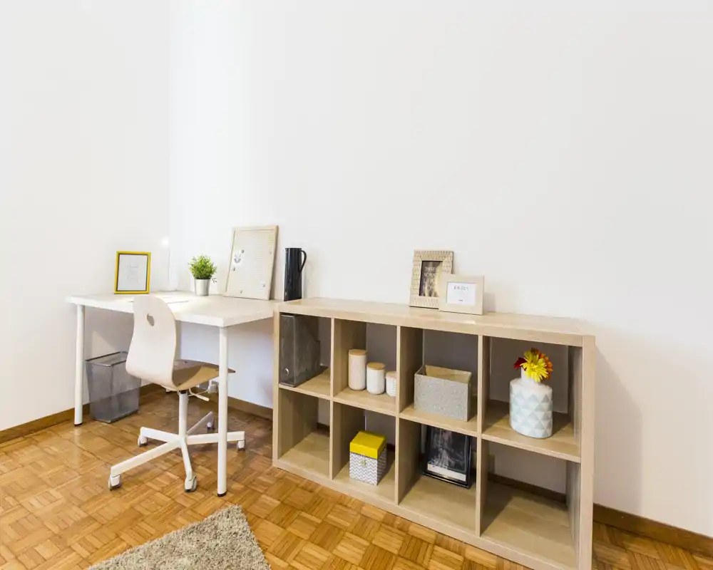 Bright, minimal living area with a workspace: white desk, swivel chair and a low cubed shelving unit with decorative boxes and a plant.