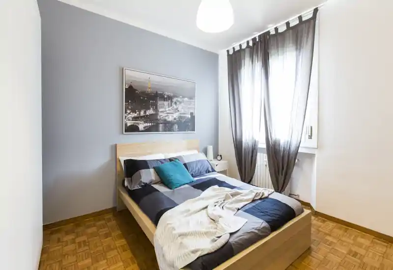 Bright, neatly styled bedroom with a wooden bed, patterned bedding and a turquoise accent pillow; natural light filters through sheer curtains.