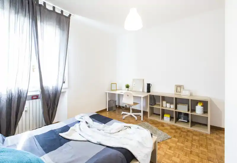 Bright, minimal bedroom with a double bed in the foreground, desk and shelving along the wall, and soft natural light from a large window.