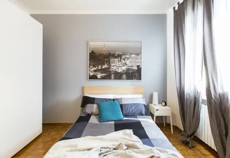 Bright, modern bedroom with a neatly made double bed, teal accent pillow and large window letting in natural light. Neutral tones and simple decor create a calm, stylish atmosphere ideal for rental listings.