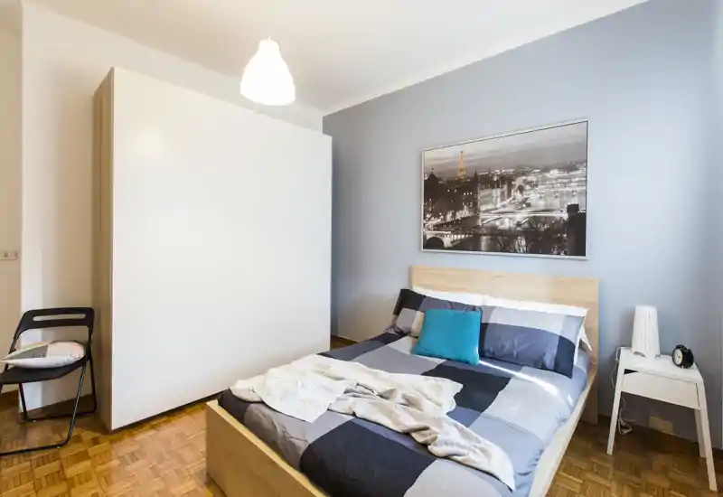 Modern, bright bedroom with a double bed, wooden headboard and a large white wardrobe. The room features clean lines, parquet flooring and a decorative city photo above the bed.