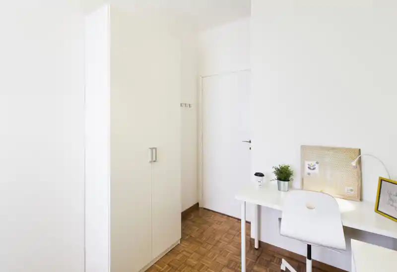 Bright, minimal bedroom corner featuring a tall wardrobe, a small desk with chair, lamp and decorative plant — ideal for a compact bedroom or study corner.