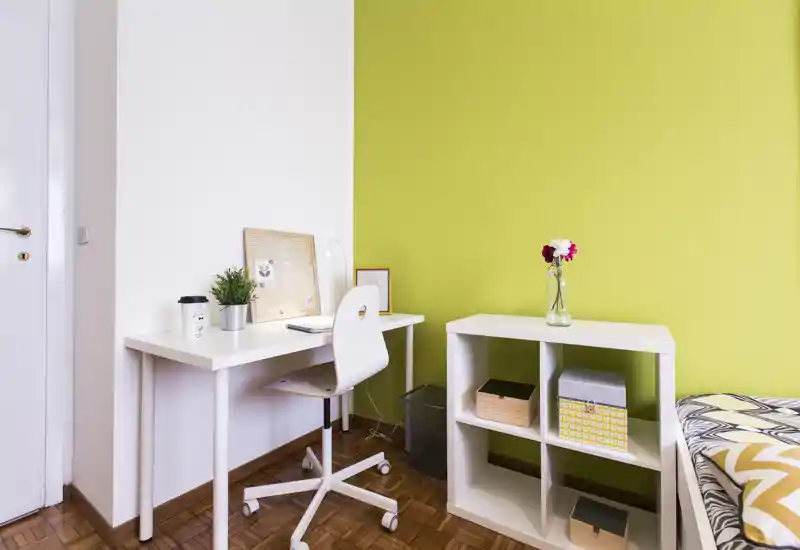 Small bedroom corner with a white study desk, swivel chair and open shelving next to a partly visible bed; bright, modern decor with a green accent wall.