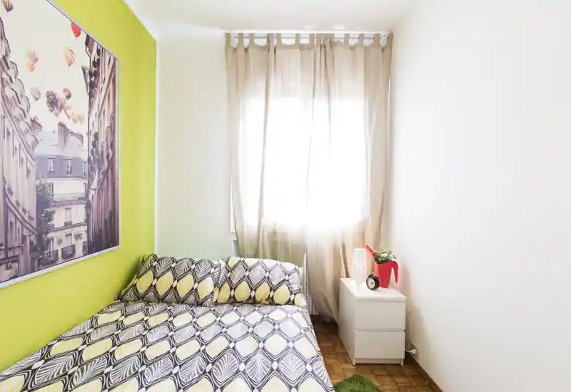 Bright, cozy single/double bedroom with patterned bedding, a green accent wall and a bedside drawer under soft natural light. Ideal for a modern, compact stay with simple decorative touches.