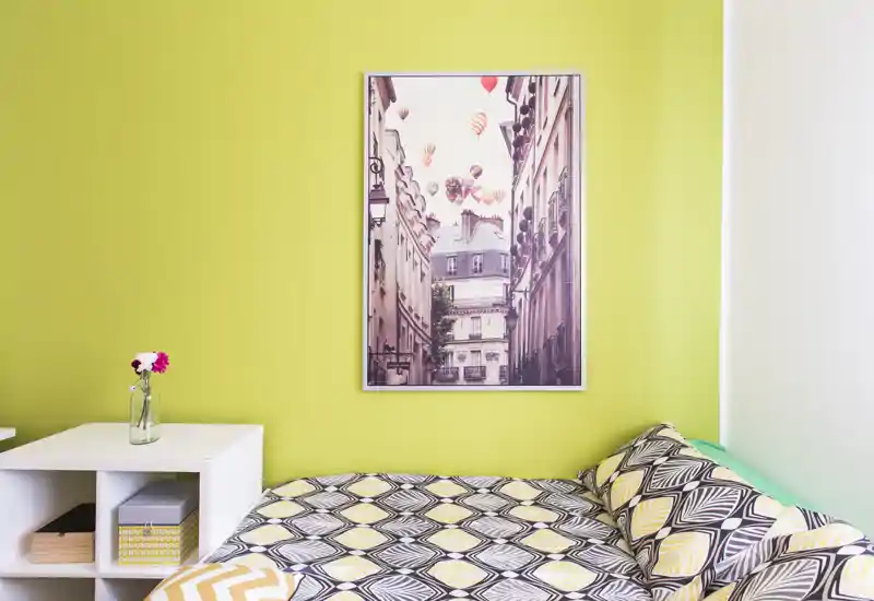 Bright, modern bedroom corner with patterned bedding, a decorative framed print on a lime wall and a small white bedside shelving unit with a vase.