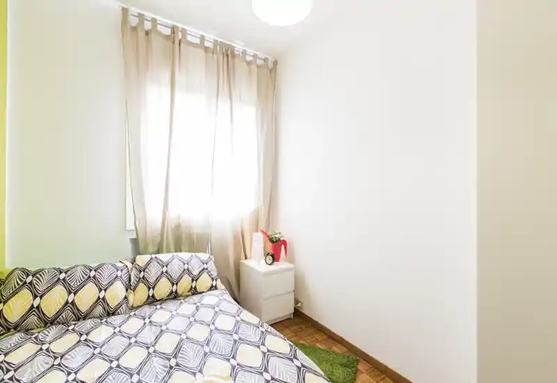 Bright, compact bedroom with patterned double bed, light curtains and a small bedside table — cozy and well-lit for a comfortable stay.