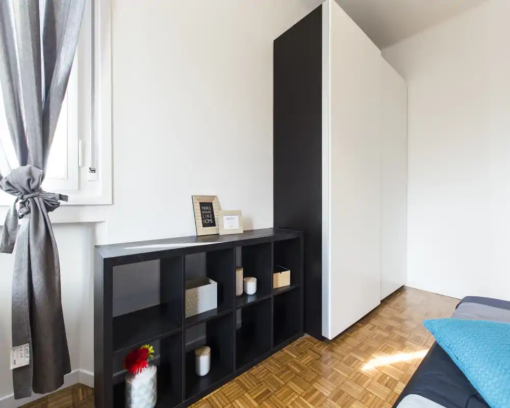 Bright, minimal bedroom corner with a small visible portion of the bed, a black shelving unit and a tall wardrobe; natural light and simple, modern decor.