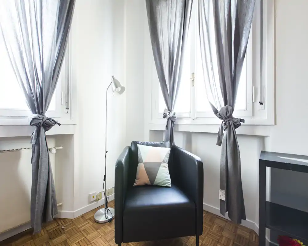 Bright, minimal living area featuring a single leather armchair by sunlit windows, a floor lamp and a decorative cushion — a cozy corner for reading or relaxing.