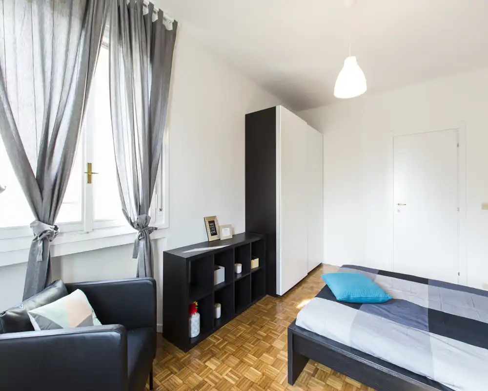 Bright, modern bedroom with a neatly made double bed, a black storage unit and a comfortable armchair by a large window letting in natural light.