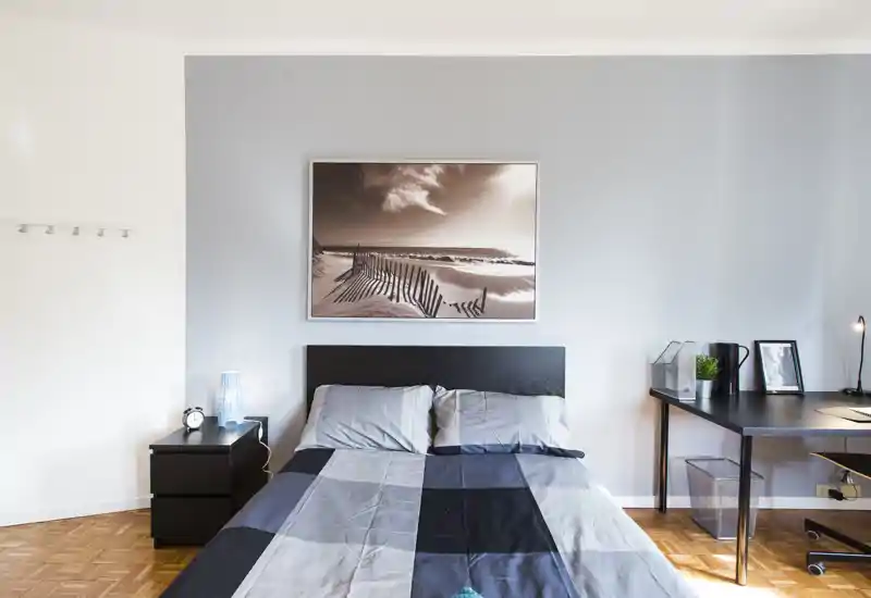 Bright, well-staged bedroom with a double bed, bedside table and a desk; calm gray tones and a framed seaside print above the headboard.