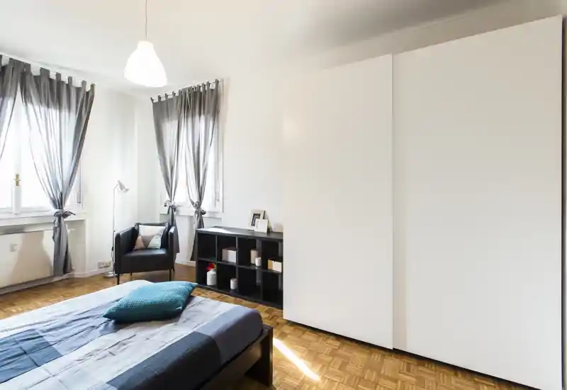 Bright, modern bedroom with a large bed, parquet floor and a minimalist white wardrobe; natural light from two windows and soft textiles create a welcoming atmosphere.