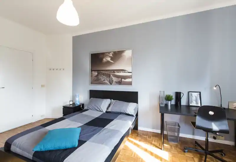 Bright, modern bedroom with a double bed, soft gray-blue color palette and a workspace desk. Natural light and minimalist decor create a calm, inviting atmosphere ideal for short-term rentals.