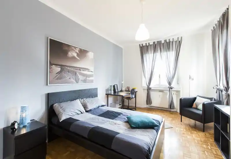 Spacious, well-lit bedroom with a double bed, bedside tables, desk area and a comfortable armchair; neutral decor and large window provide airy natural light.