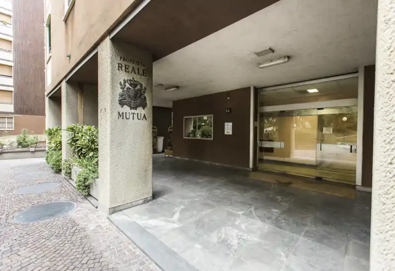 Covered building entrance with sliding glass doors, tiled driveway and a sheltered portico — clear view of the main access and intercom area.