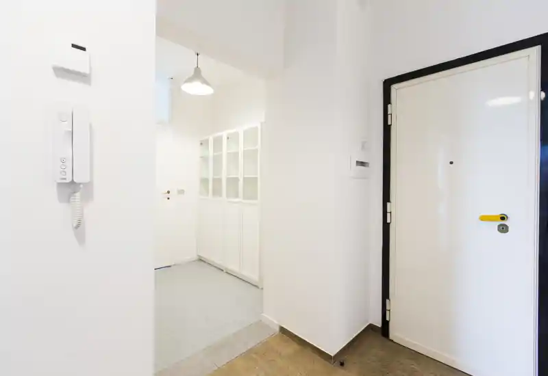 Bright, minimal entrance hall with a white security door, wall-mounted intercom and built-in storage visible through the doorway.