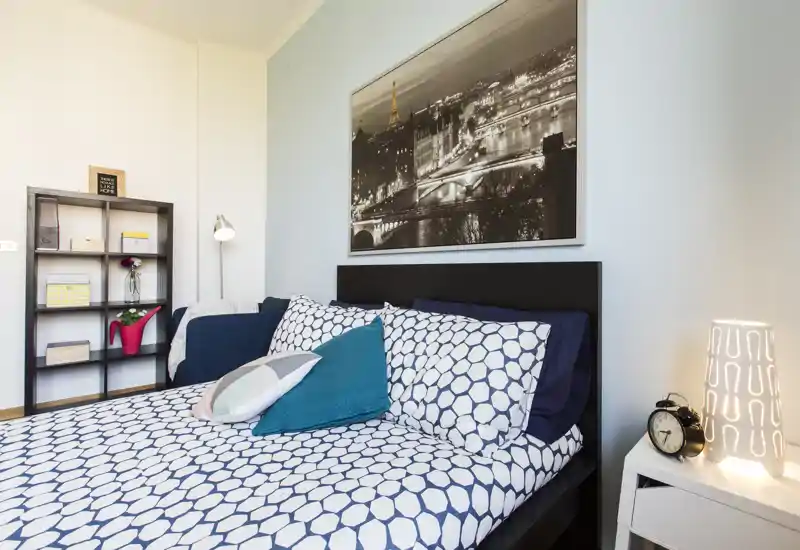 Bright, well-styled bedroom with a patterned bedspread, decorative cushions and bedside lamp. The large framed cityscape above the headboard and tidy shelving add modern character.