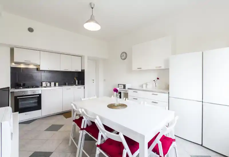 Bright, modern kitchen with integrated oven, ample white cabinetry and a central dining table with red-cushioned chairs — a clean and inviting space for meals and cooking.